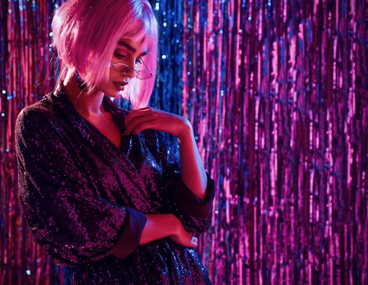 glamorous girl in a stylish brilliant evening dress, stands thoughtful in the light of neons in a di...