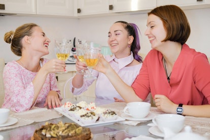 Three women toast their wine glasses while laughing in a bright kitchen.