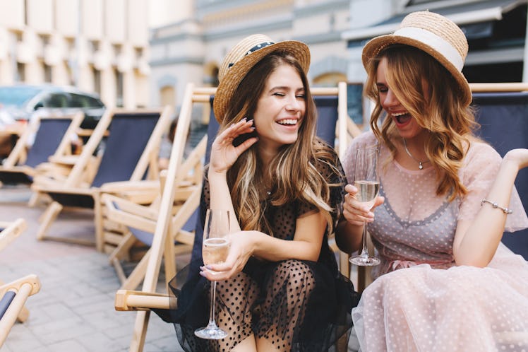 Two girls in dresses and hats laugh and hold their champagne glasses on a patio.