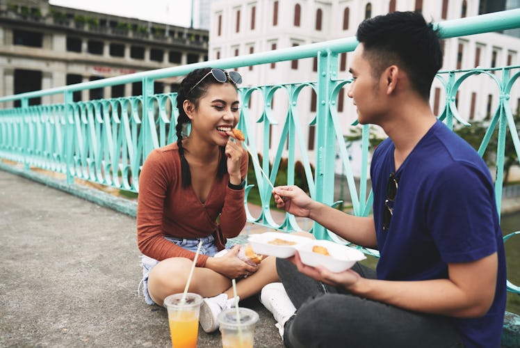 Young man feeding his girlfriend while they eating fast food on the ground in the city