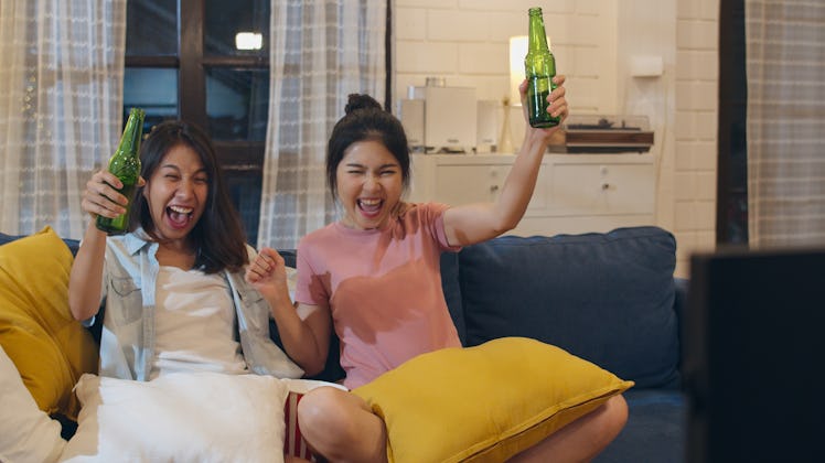 Two female friends cheer and raise their beer bottles on the couch while watching a game.