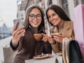 Two brunette girls hold their coffee cups and snap a selfie at an outdoor café.