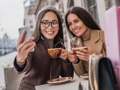 Two brunette girls hold their coffee cups and snap a selfie at an outdoor café.