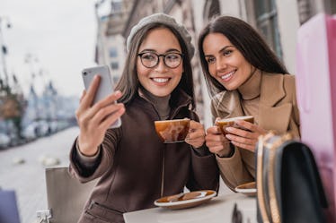 Two brunette girls hold their coffee cups and snap a selfie at an outdoor café.