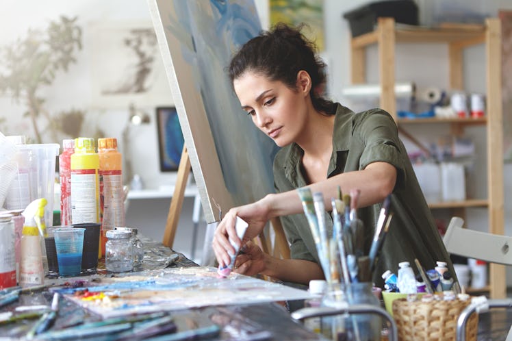 Picture of serious concentrated young Caucasian female artist sitting at desk with painting accessor...