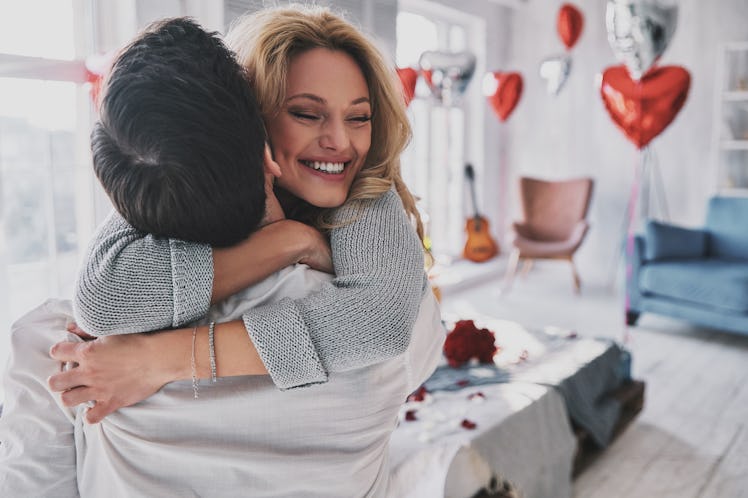 A happy blonde woman hugs her boyfriend who is lifting her up in the bedroom on Valentine's Day.