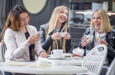 Three girls laugh while enjoying tea and macarons at an outdoor café.