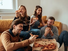 Two couples laugh and enjoy pizza in a bright living room.