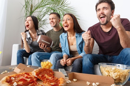 A group of four friends watch a football game on TV with a box of pizza and snacks in front of them.