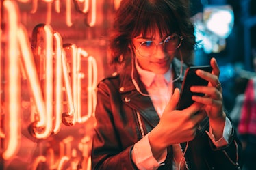 Young stylish female hipster standing near neon red light in night city and listening to music in he...