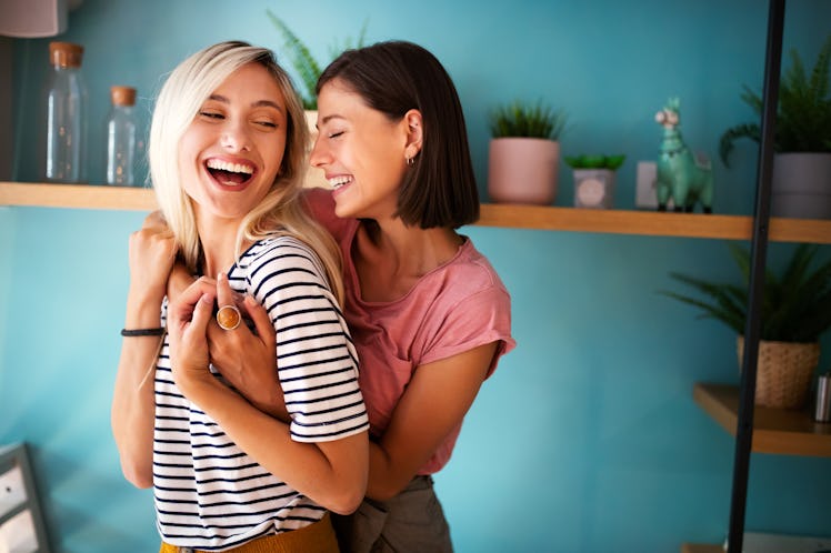 Two women smile and embrace in a blue room on a sunny day.