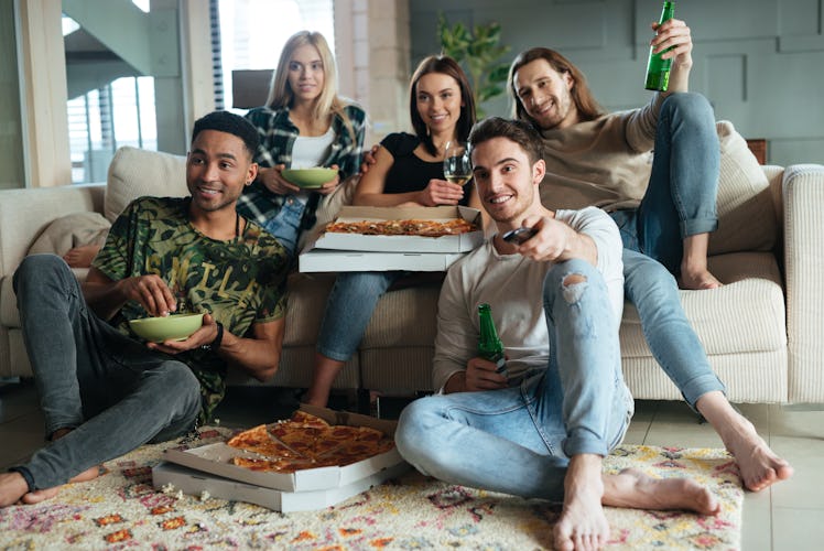 Five friends sit in a living room while watching TV and enjoying beer and pizza.