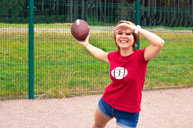 A blonde-haired woman in a red football shirt prepares to throw a football outside in the park. 