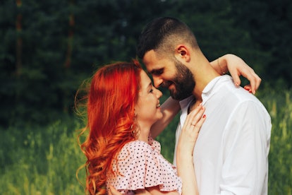 A woman with red hair hugs her boyfriend and smiles in a sunny field.