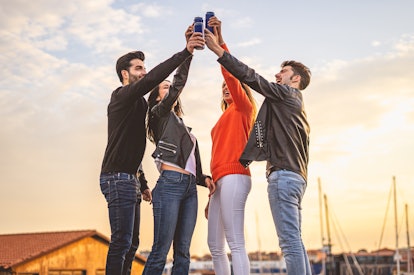 A group of friends toast their beer cans on a rooftop at sunset.