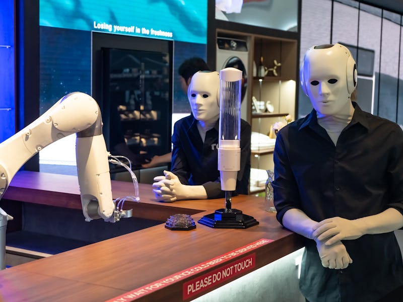 Robots in human form stand behind the bar, where another robotic arm pours them drinks. People in su...