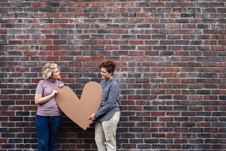 A happy couple holds up a large cardboard heart in front of a brick wall.