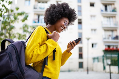 A young woman wearing a backpack and glasses is walking through the city and smiling at her phone. S...