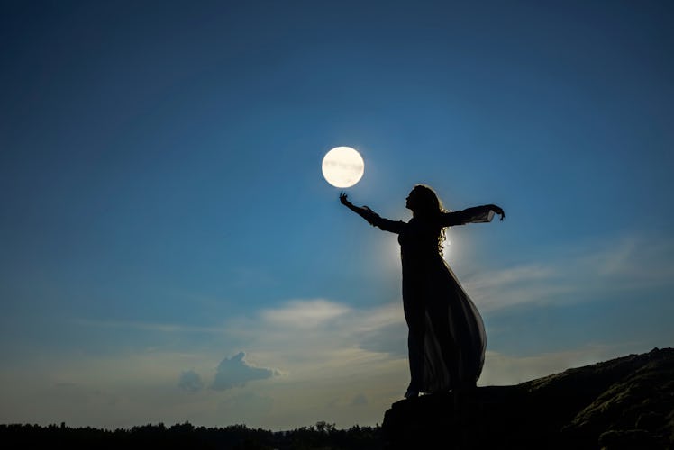 silhoutee of woman standing on top of mountain. female hold in hand full moon above forest trees. ba...