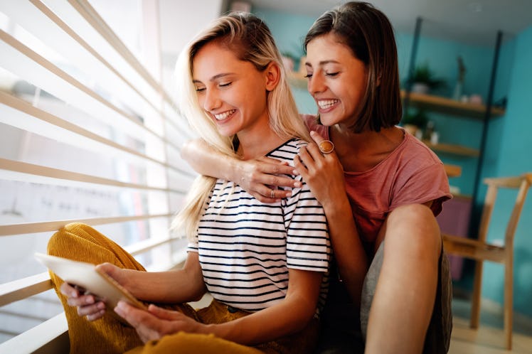 A happy couple sits on the floor and reads a card next to a sunny window.