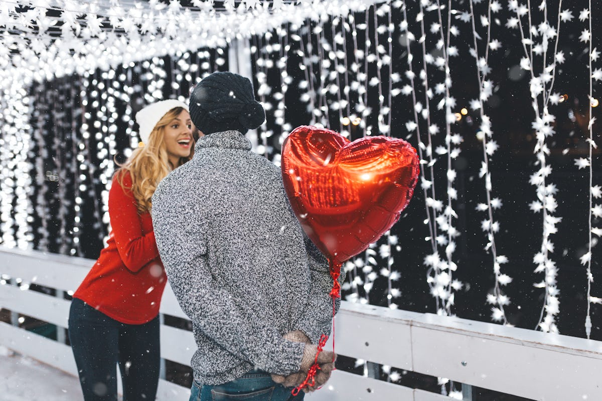 A woman smiles at her boyfriend who's holding a heart-shaped balloon behind his back on the ice rink.