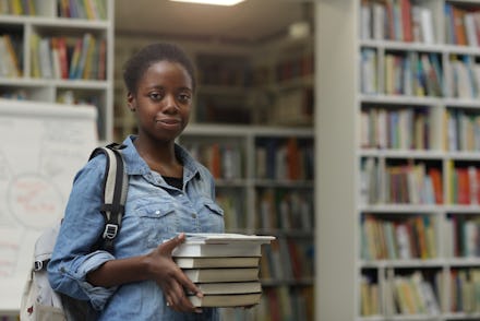 Portrait of African young woman looking at camera while holding heap of books in her hands in the li...