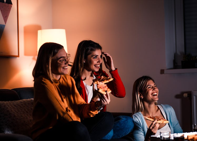 Three female friends laugh, eat pizza, and watch TV in a dimly-lit living room.