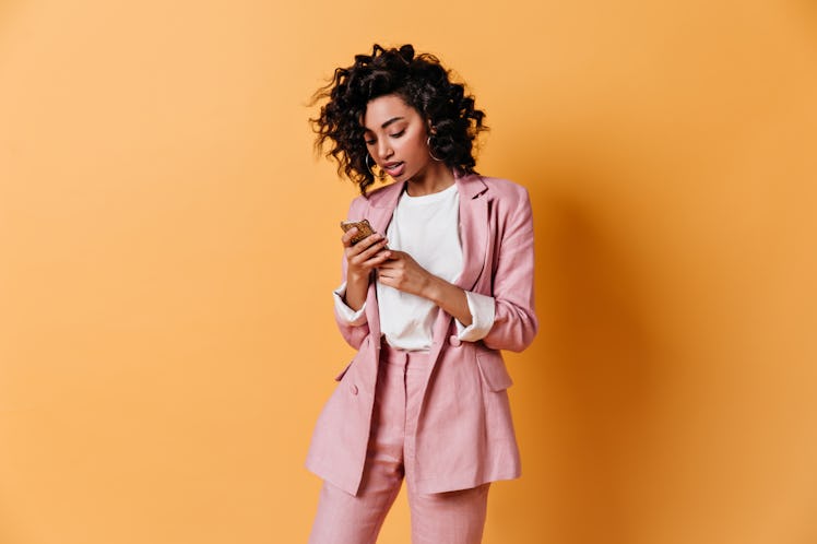 Focused curly woman texting message. Studio shot of elegant girl holding smartphone on yellow backgr...
