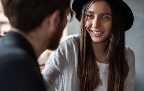 Cheerful young woman in hat smiling and talking with anonymous man during romantic date in cafe