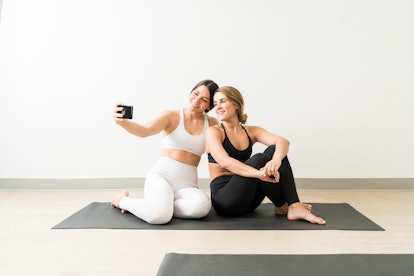 Two happy friends in workout clothes pose for a selfie on a yoga mat.