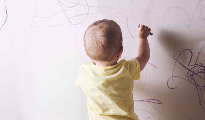 Baby boy drawing with wax crayon on plasterboard wall. He is with his back towards