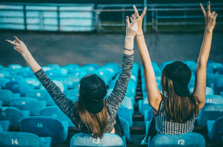 Two women wearing backwards baseball caps cheer on their team in the bleachers.
