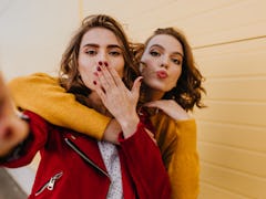 Two women blow a kiss at the camera while posing for a selfie outside.