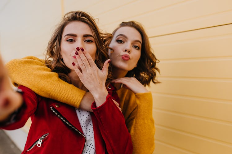 Two women blow a kiss at the camera while posing for a selfie outside.