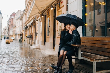 A girl smiles and sits on her boyfriend's lap under an umbrella on a rainy afternoon in a city.