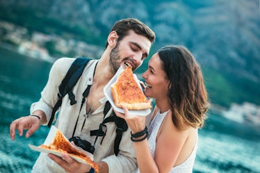 A happy couple who's traveling enjoys a slice of pizza by the water.