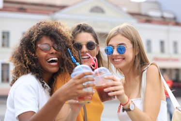 Three trendy girls in sunglasses toast their smoothies and smile on a sunny day in the city.