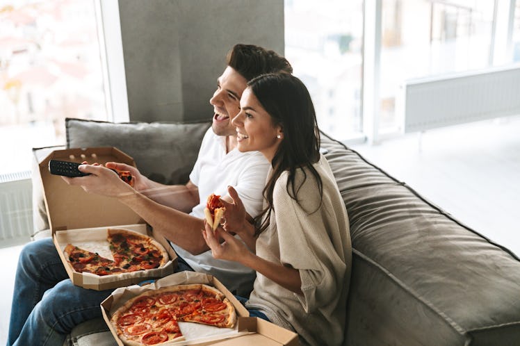 A young couple enjoys pizza on the couch and watches TV.