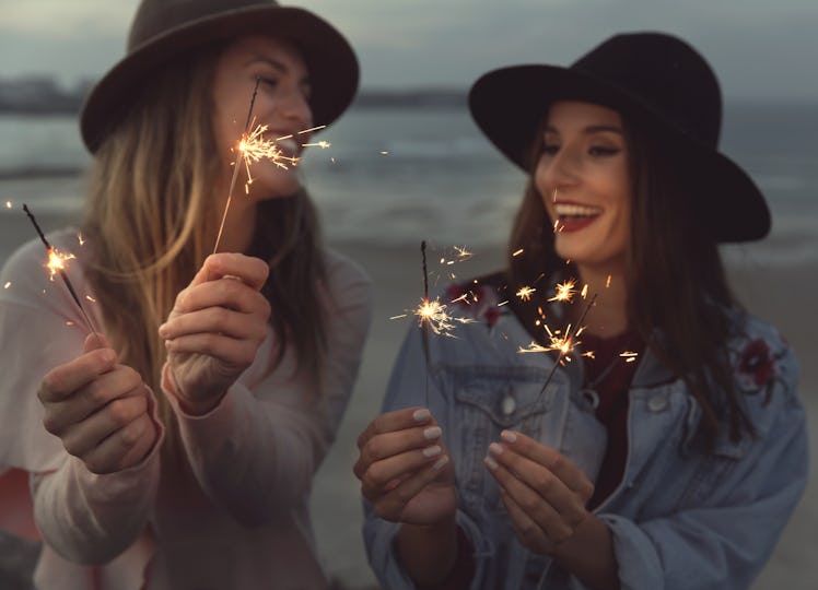 Two best friends in felt hats laugh and hold sparklers on the beach.