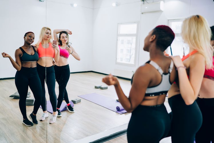A group of friends in workout pants, sneakers, and sports bras pose for a mirror selfie at the gym.