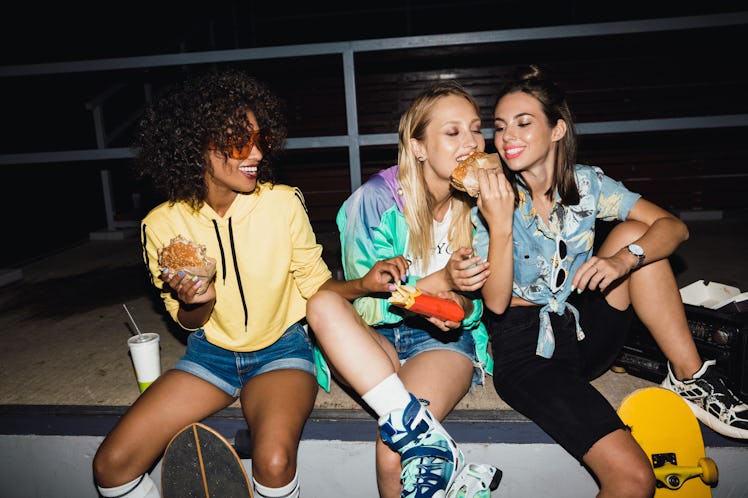 A group of three friends at night sit on a sidewalk and enjoy fries and burgers.