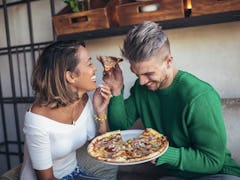 A couple laughs and eats pizza on a couch in a modern living room.