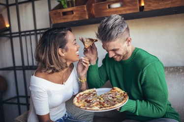 A couple laughs and eats pizza on a couch in a modern living room.