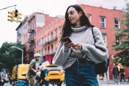 Beautiful slender woman in glasses with black bag wearing denim and sweater looking away while brows...