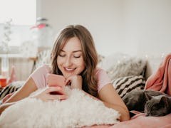 A happy woman looks at her phone with her cat sleeping next to her. 