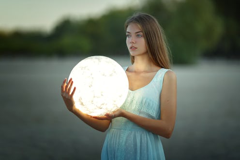 Attractive young girl in a blue dress sits on a sandy beach and holds the moon in her hands