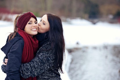A woman hugs her girlfriend and gives her a kiss on the cheek outside in the snow.