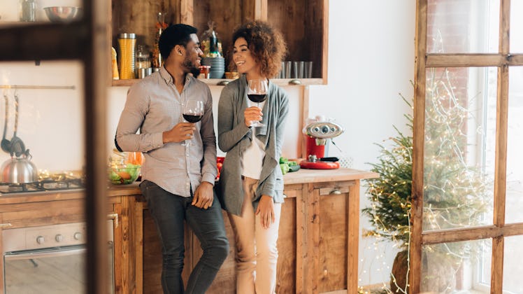 A couple laughs while holding their wine glasses in the kitchen on a sunny day.