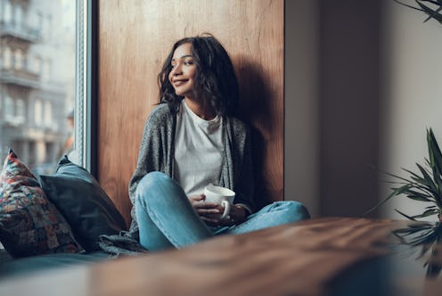 Calm relaxed young woman looking outdoors and smiling while sitting on the window sill with a cup of...