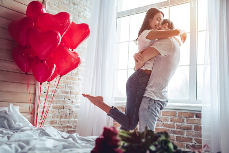 A happy couple embraces in a bedroom with heart-shaped balloons next to them on Valentine's Day.